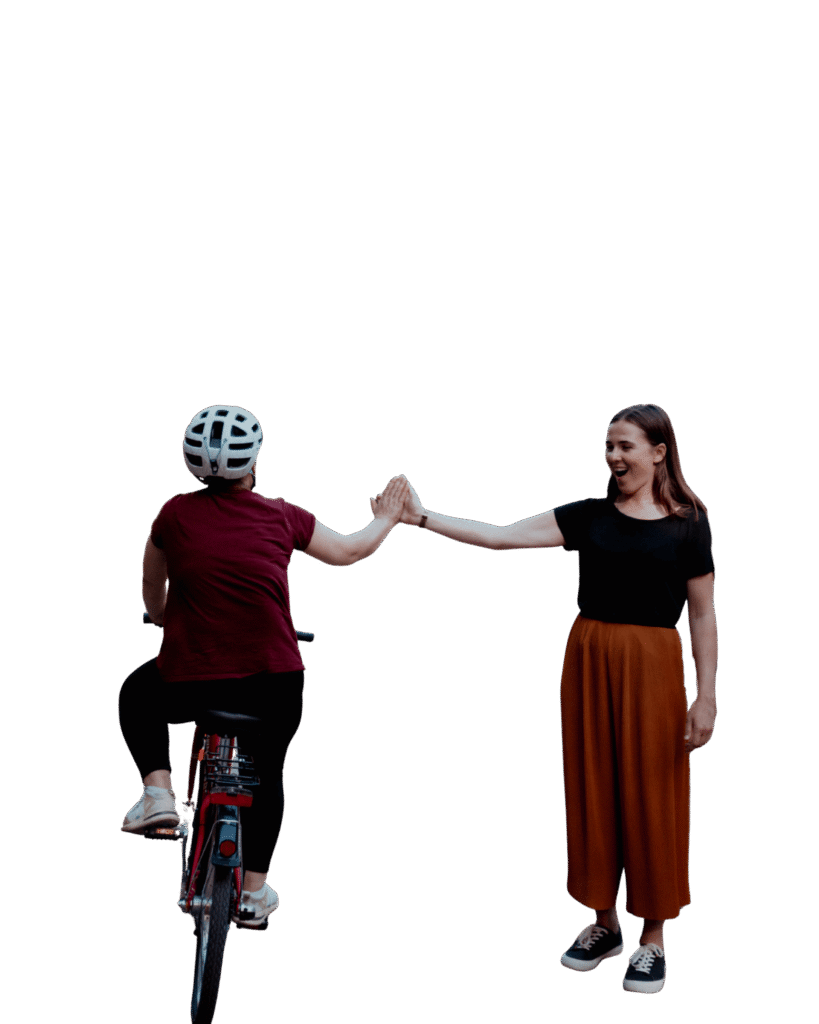 Frau auf einem Fahrrad gibt einer lachenden Frau am Wegesrand ein High-Five in einem Park.