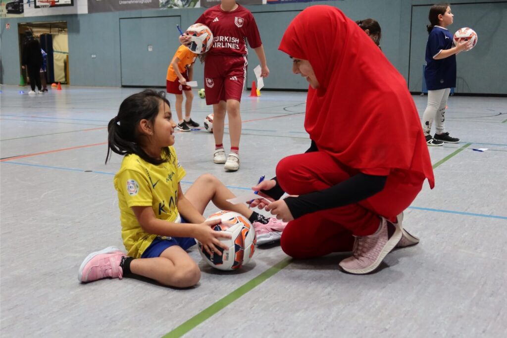 Trainerin im roten Sportoutfit spricht in einer Turnhalle mit einem sitzenden Mädchen im gelben Trikot, das einen Fußball hält.