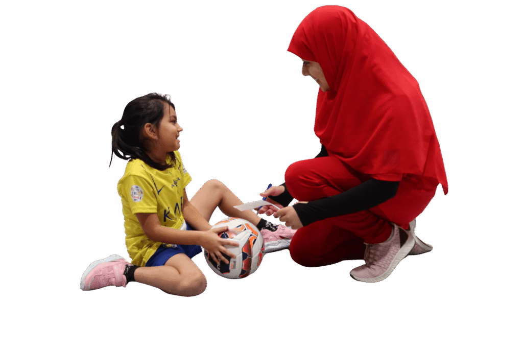 Trainerin im roten Sportoutfit spricht in einer Turnhalle mit einem sitzenden Mädchen im gelben Trikot, das einen Fußball hält.