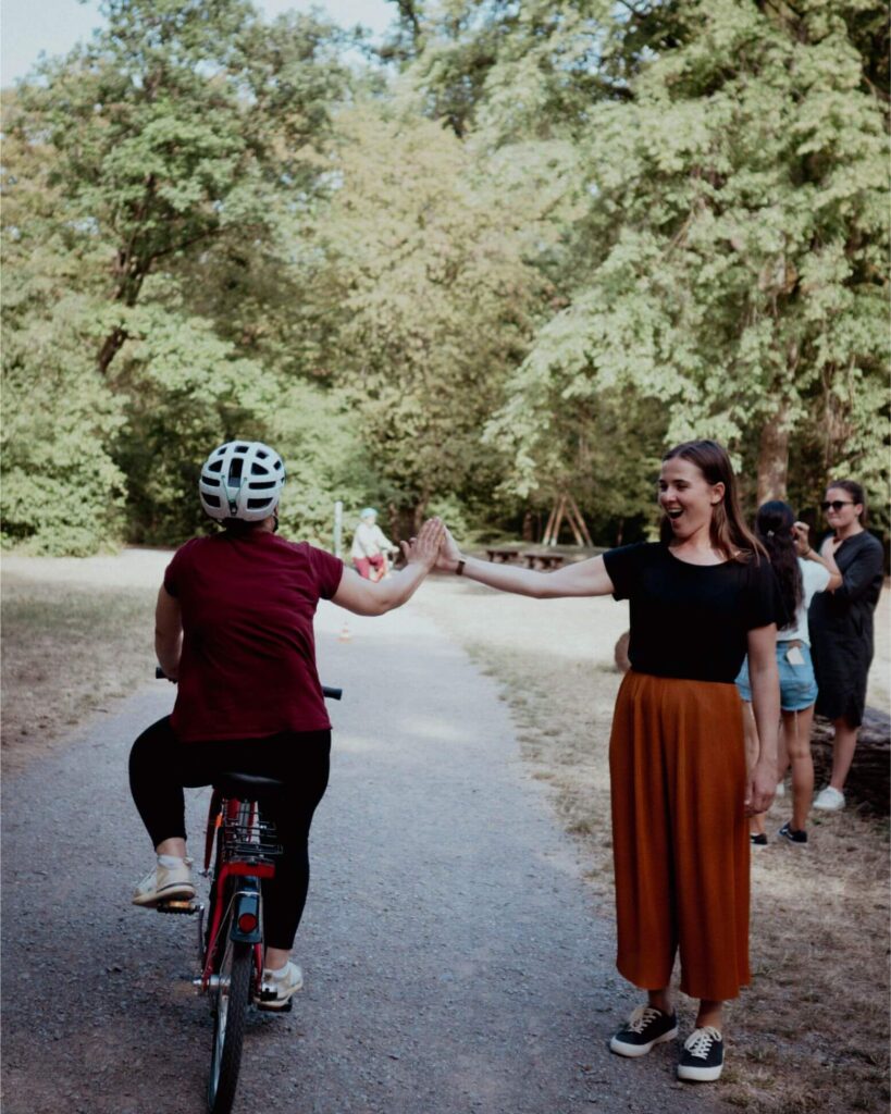 Frau auf einem Fahrrad gibt einer lachenden Frau am Wegesrand ein High-Five in einem Park.